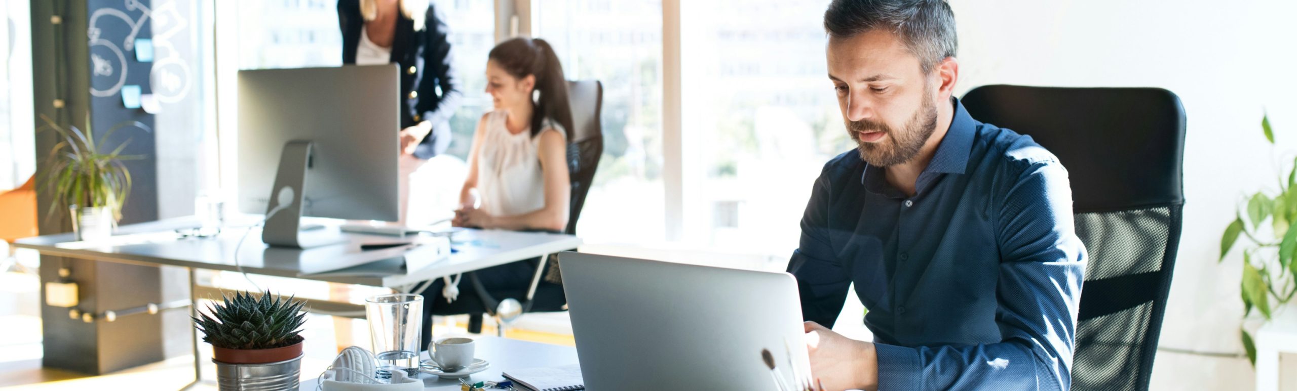 Im Vordergrund arbeitet ein Mitarbeiter am Laptop, im Hintergrund ist ein Großraumbüro, in dem sich zwei Mitarbeiter am Schreibtisch unterhalten.